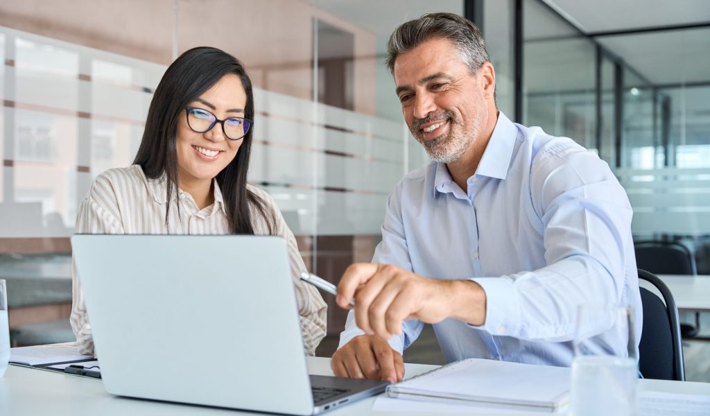 Mid aged Latin male manager mentor teaching young Asian female worker looking at laptop discussing corporate strategy in teamwork, working on computer in office at international team meeting.